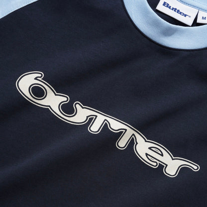 Close-up of a Butter raglan tee in navy blue with a light blue ribbed collar and a large white embroidered logo outlined in black on the chest.